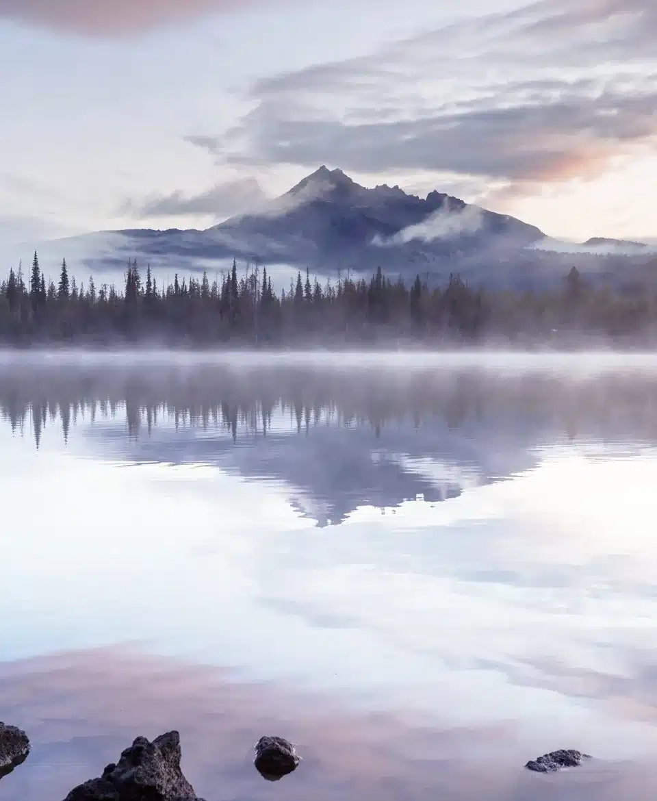 A still lake at dawn, mist rising, mountains reflected in the water